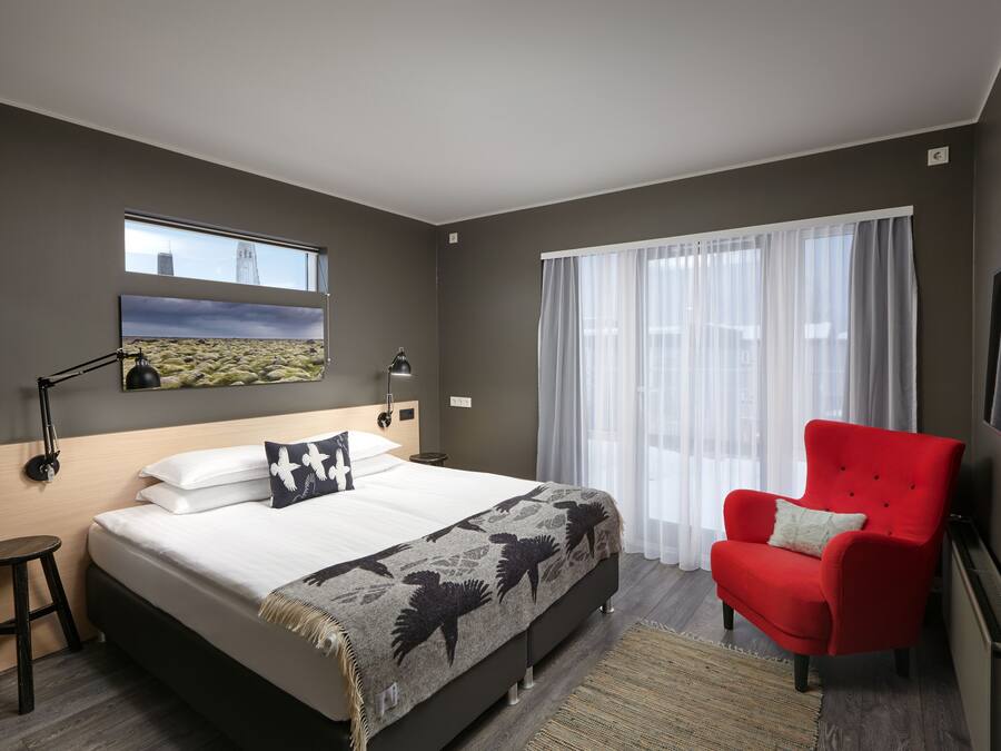 Quarto de hotel moderno com uma cama grande, uma poltrona vermelha, uma janela com vista para a cidade e uma televisão na parede.