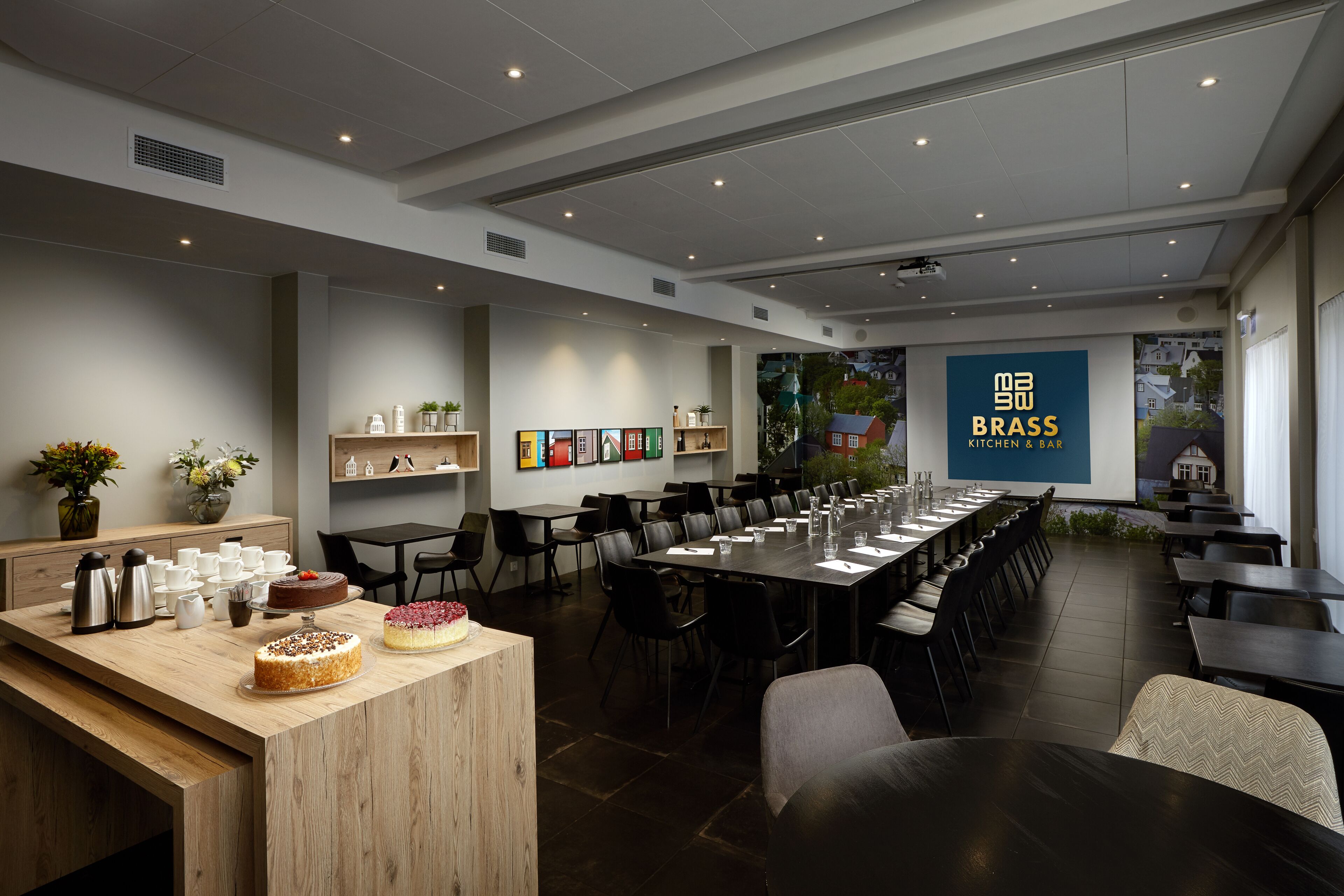 Un restaurante moderno con una larga mesa compartida, una barra de madera con pasteles y un gran mural de casas.