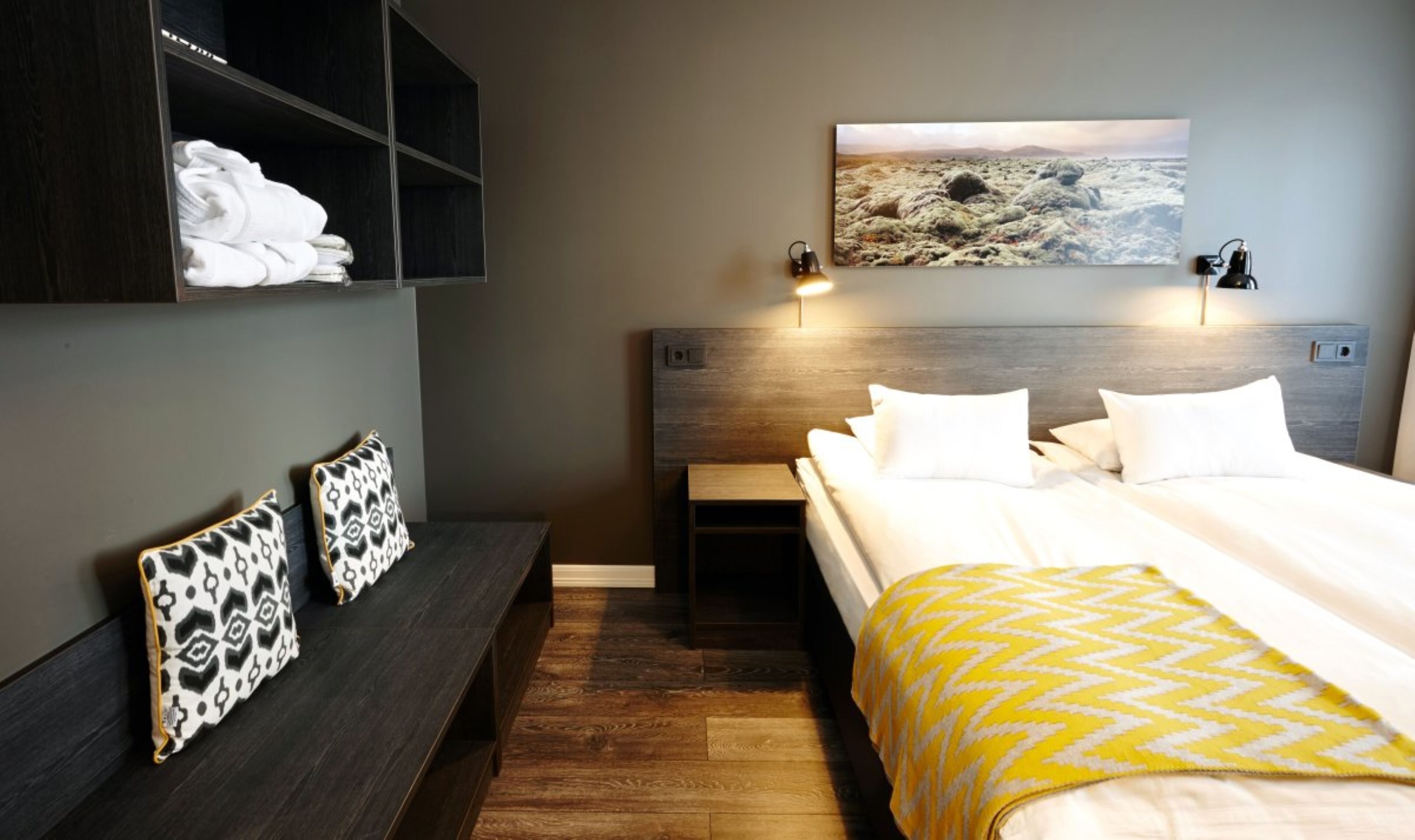 A hotel room with a bed, bedside tables, a bench with pillows, a wall-mounted painting, and a nightstand with a lamp.