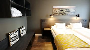 A hotel room with a bed, bedside tables, a bench with pillows, a wall-mounted painting, and a nightstand with a lamp.