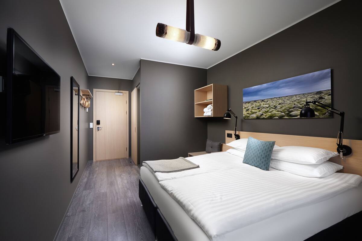 A modern hotel room with a large bed, a mounted TV, a painting, and a desk with a lamp.
