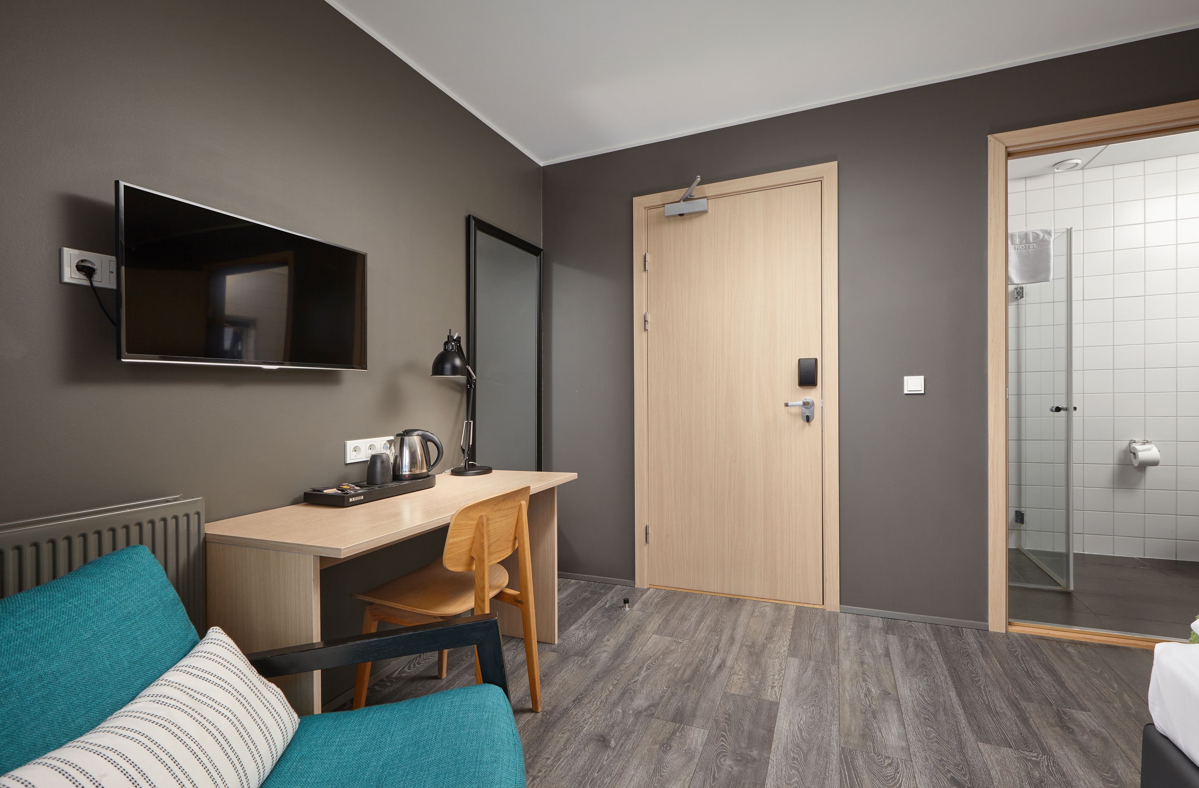Quarto de hotel moderno com escrivaninha, cadeira e um espelho grande. É visível uma porta de madeira e uma área de chuveiro.
