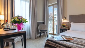 A hotel room with a bed, a desk with a vase of flowers, a chair, and a view of a building through the window. - Fragrance Hotel St. Peter (Rome)