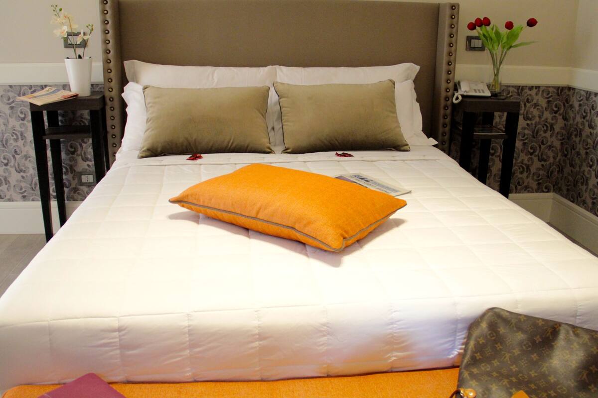 A neatly made bed with white bedding, two pillows, and an orange accent pillow.