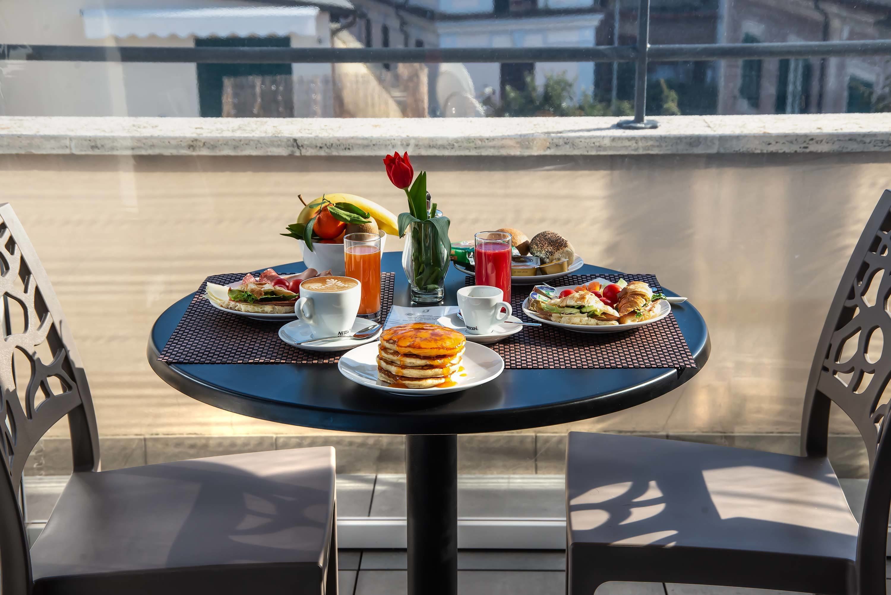 A round table with breakfast items including pancakes, fruit, and beverages, set on a balcony with a view of buildings.