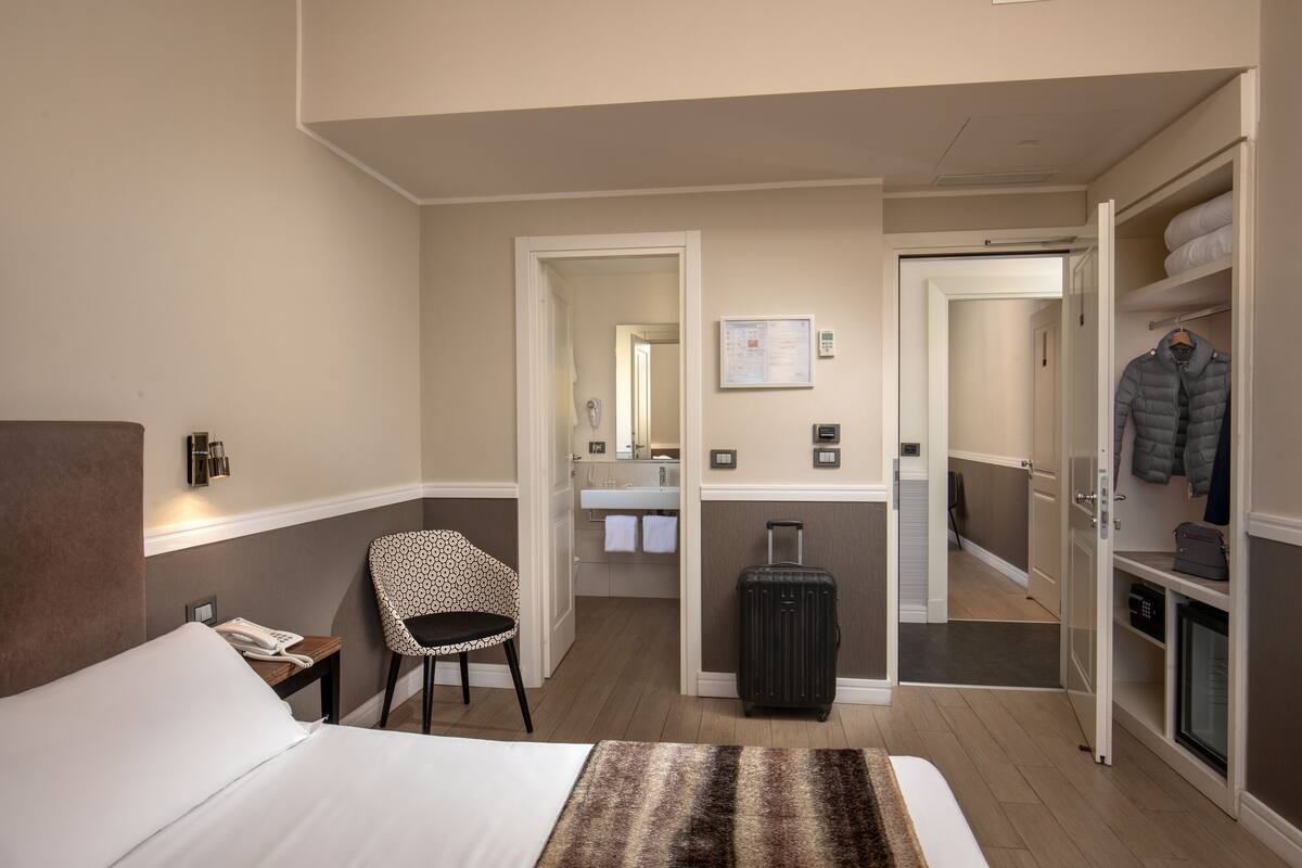 A hotel room with a bed, a chair, a desk, a suitcase, and a wardrobe.