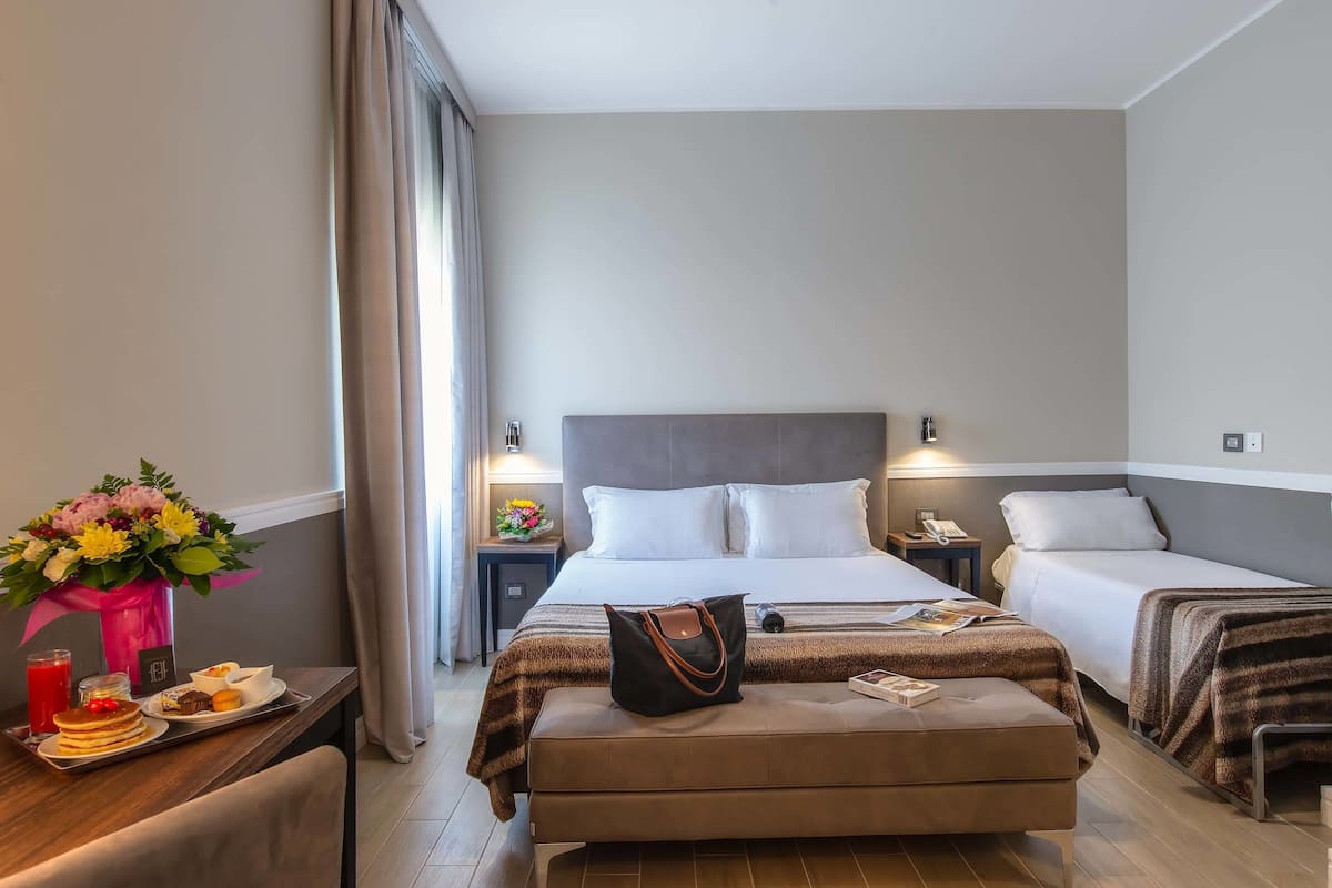 A hotel room with a large bed, two smaller beds, a desk with breakfast, a vase of flowers, and a nightstand with a lamp.