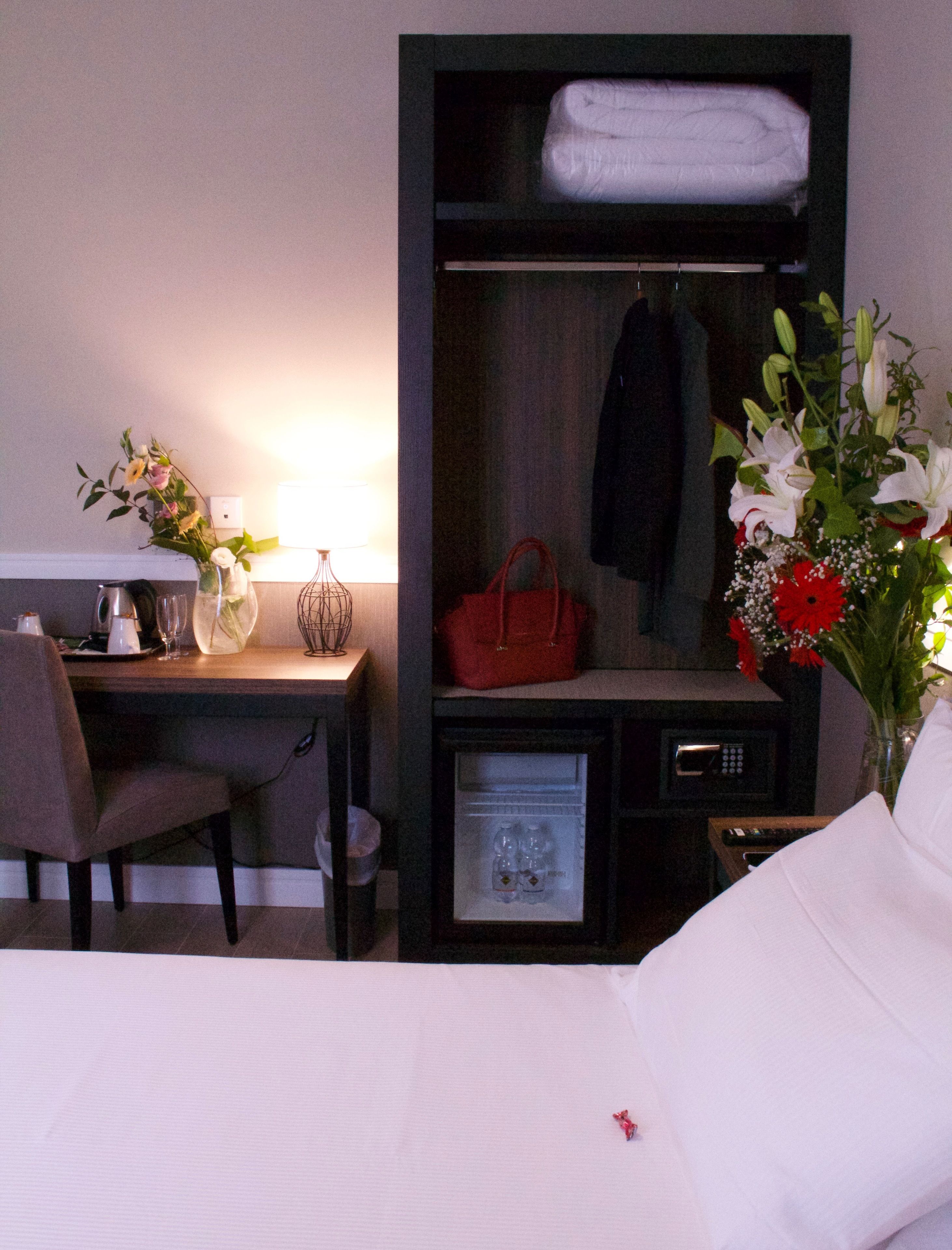 Ein Hotelzimmer mit einem Bett, einem Schreibtisch mit Lampe, einem kleinen Kühlschrank, einem Kleiderständer und einer Vase mit Blumen.