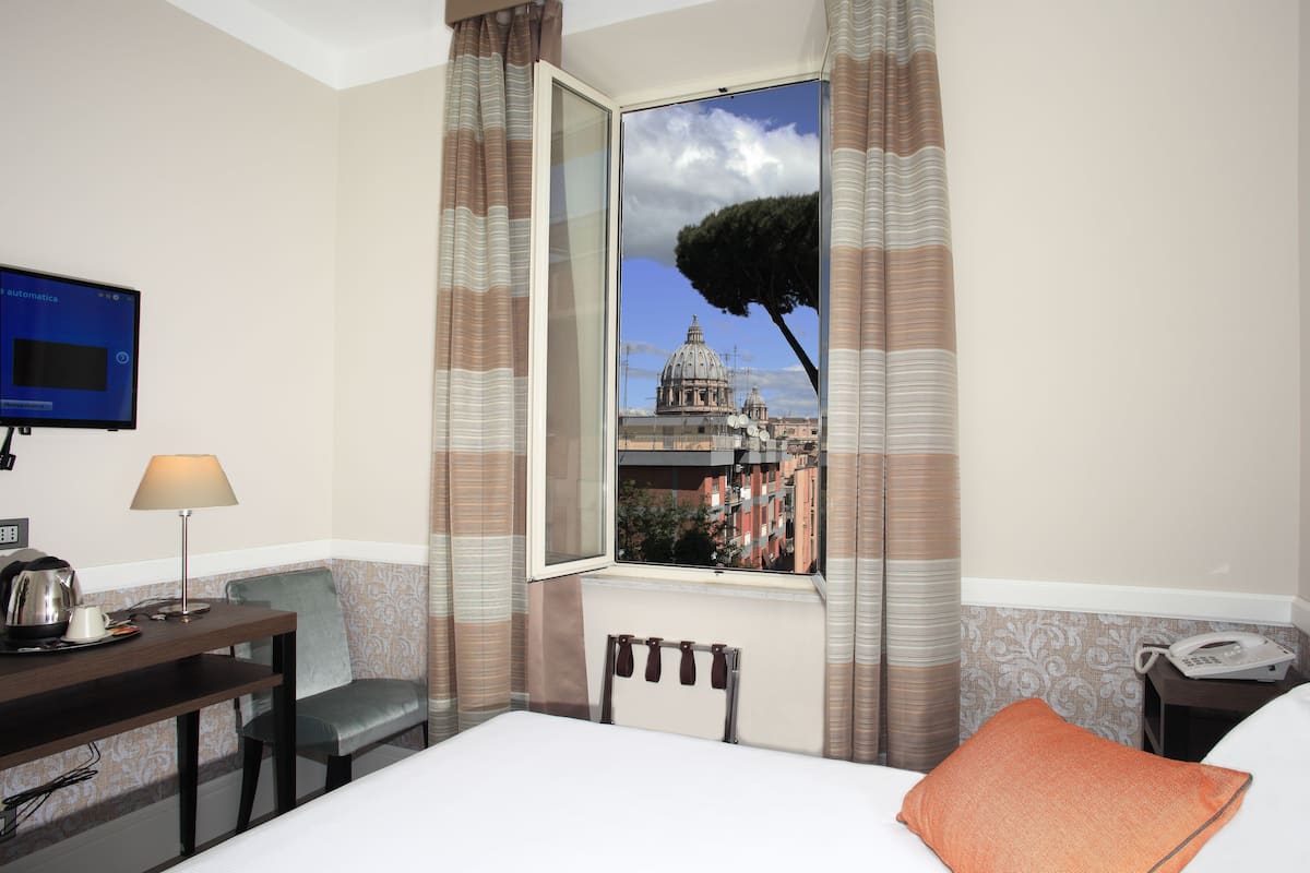 A hotel room with a bed, a desk with a lamp, a chair, a TV, and a window with a view of a building.