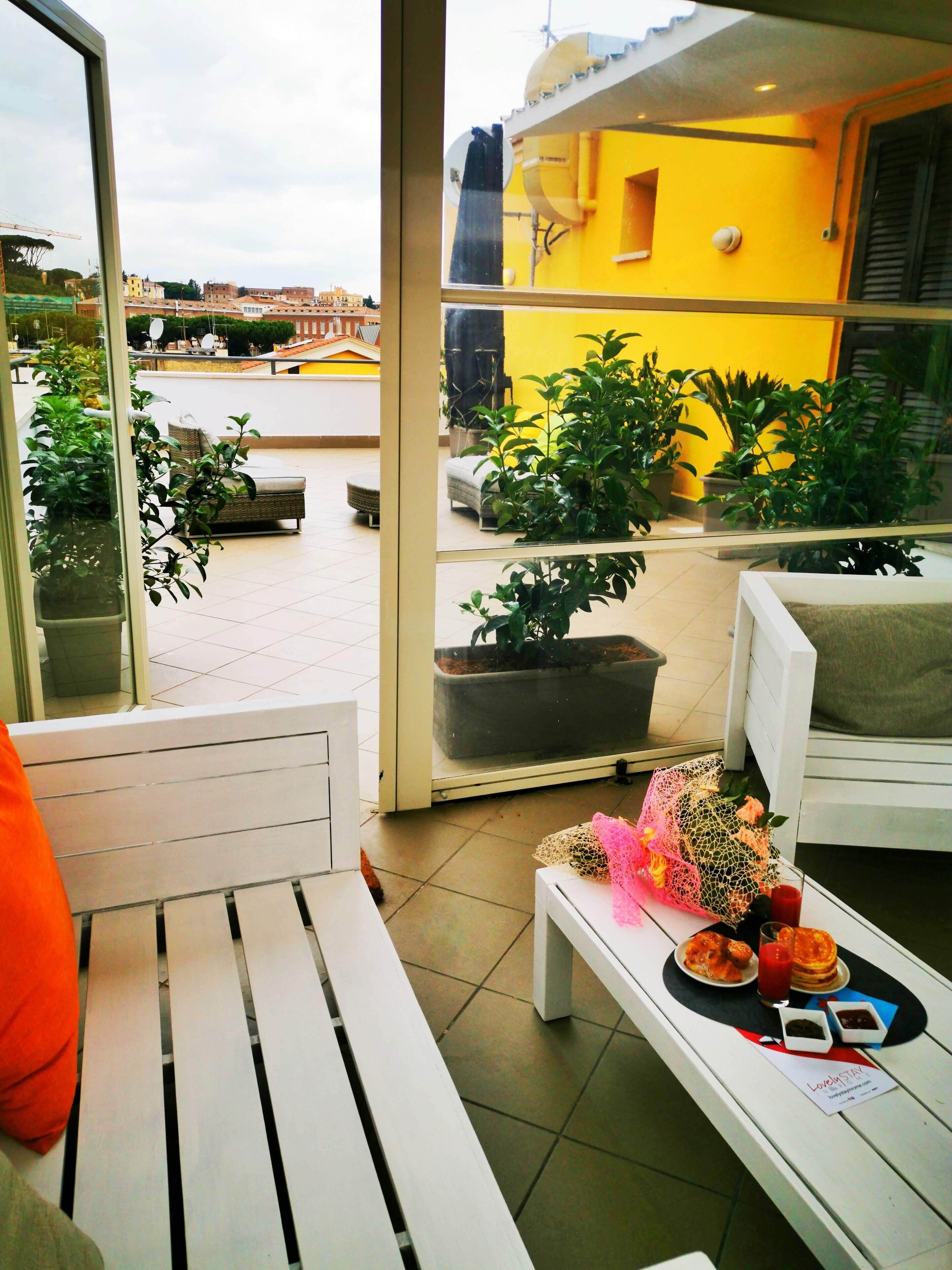 A balcony with a white bench, potted plants, and a table with a meal and a glass of red drink.