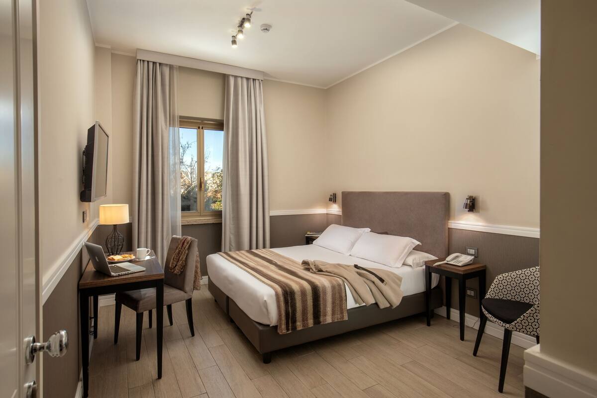 A hotel room with a bed, a desk with a laptop, a chair, a bedside table with a lamp, and a window with curtains.