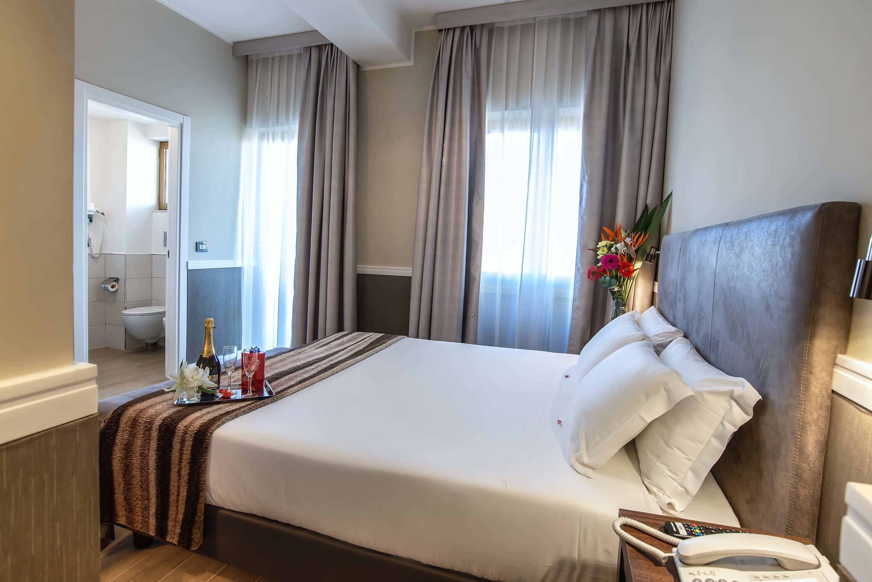 A hotel room with a large bed, a bedside phone, and a tray with a bottle and glasses.