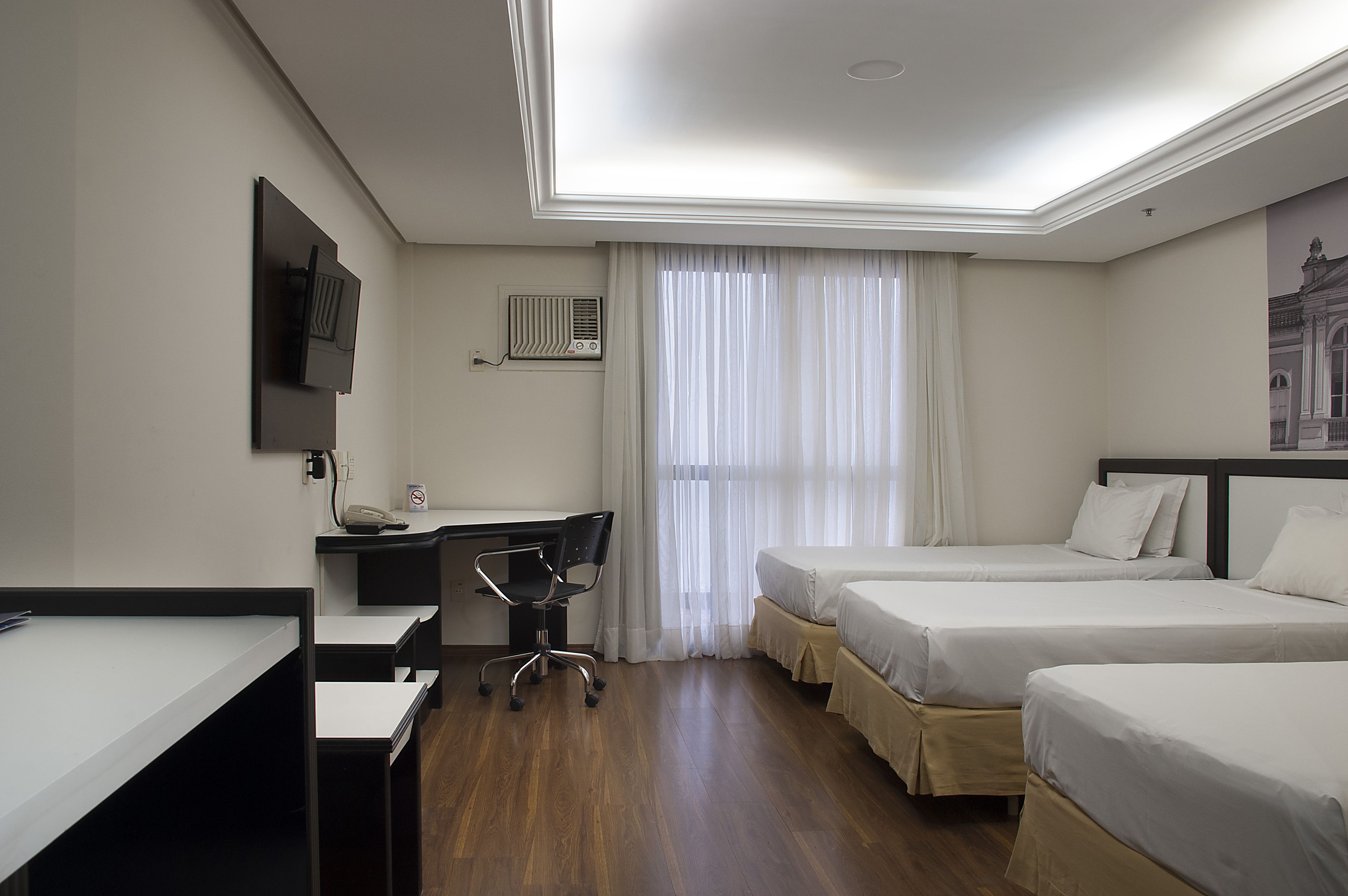 A hotel room with a desk, two beds, a chair, a TV, and a window with curtains.