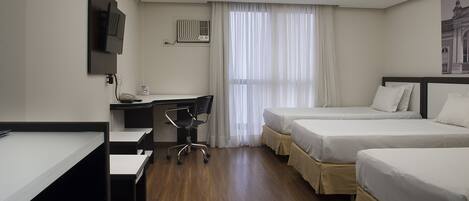 A hotel room with a desk, two beds, a chair, a TV, and a window with curtains.