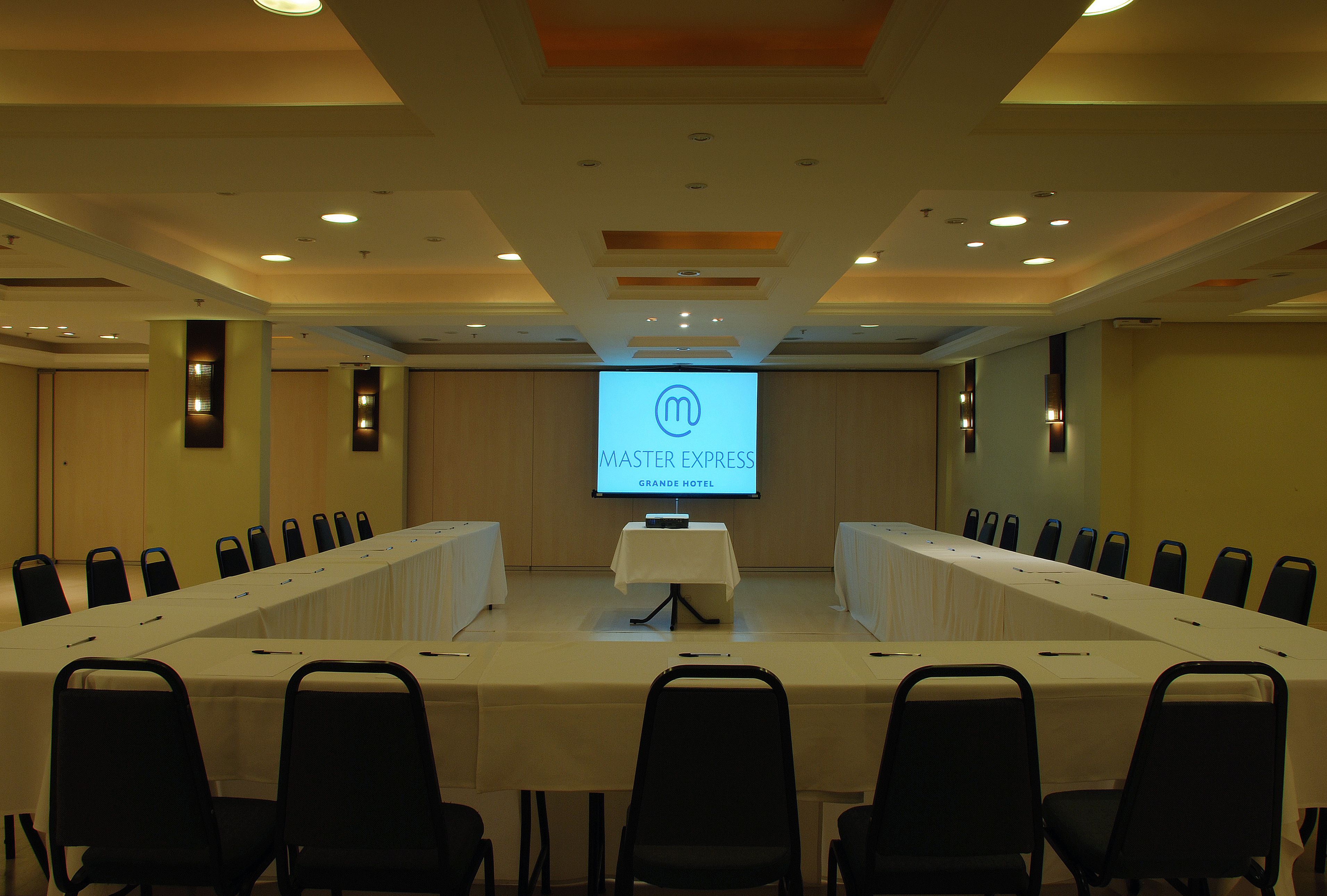 A conference room with a long table, multiple chairs, and a screen displaying 'Master Express Grande Hotel'.