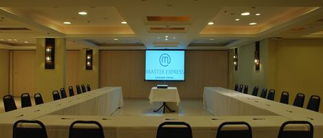 A conference room with a long table, multiple chairs, and a screen displaying 'Master Express Grande Hotel'.