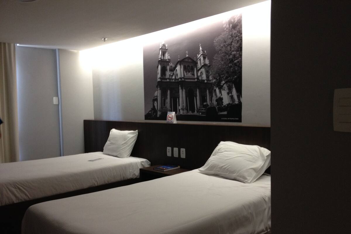 A hotel room with two single beds, a nightstand with a book, a wall-mounted light, and a framed picture of a building.