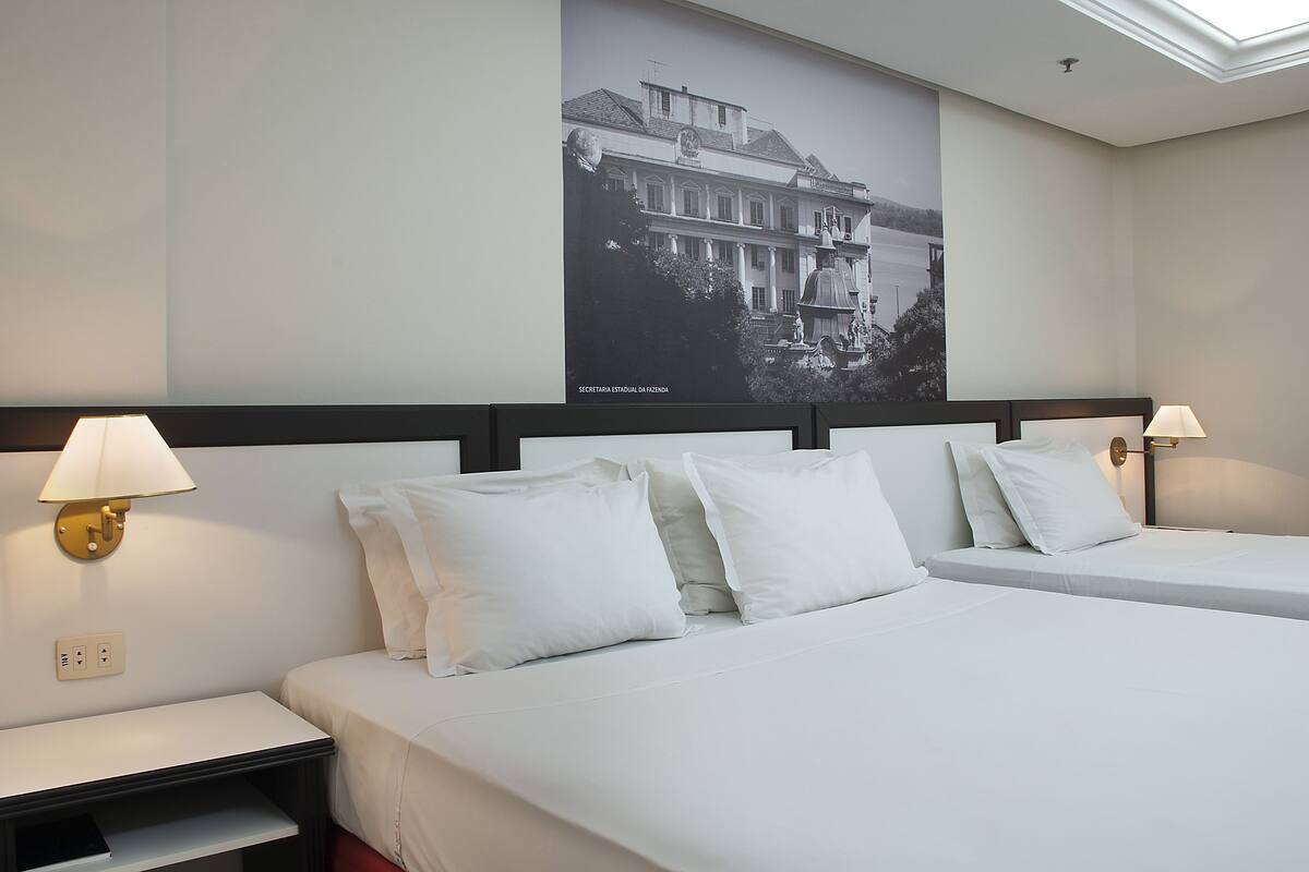 A hotel room with two beds, a nightstand, a wall-mounted lamp, and a large poster of a building.
