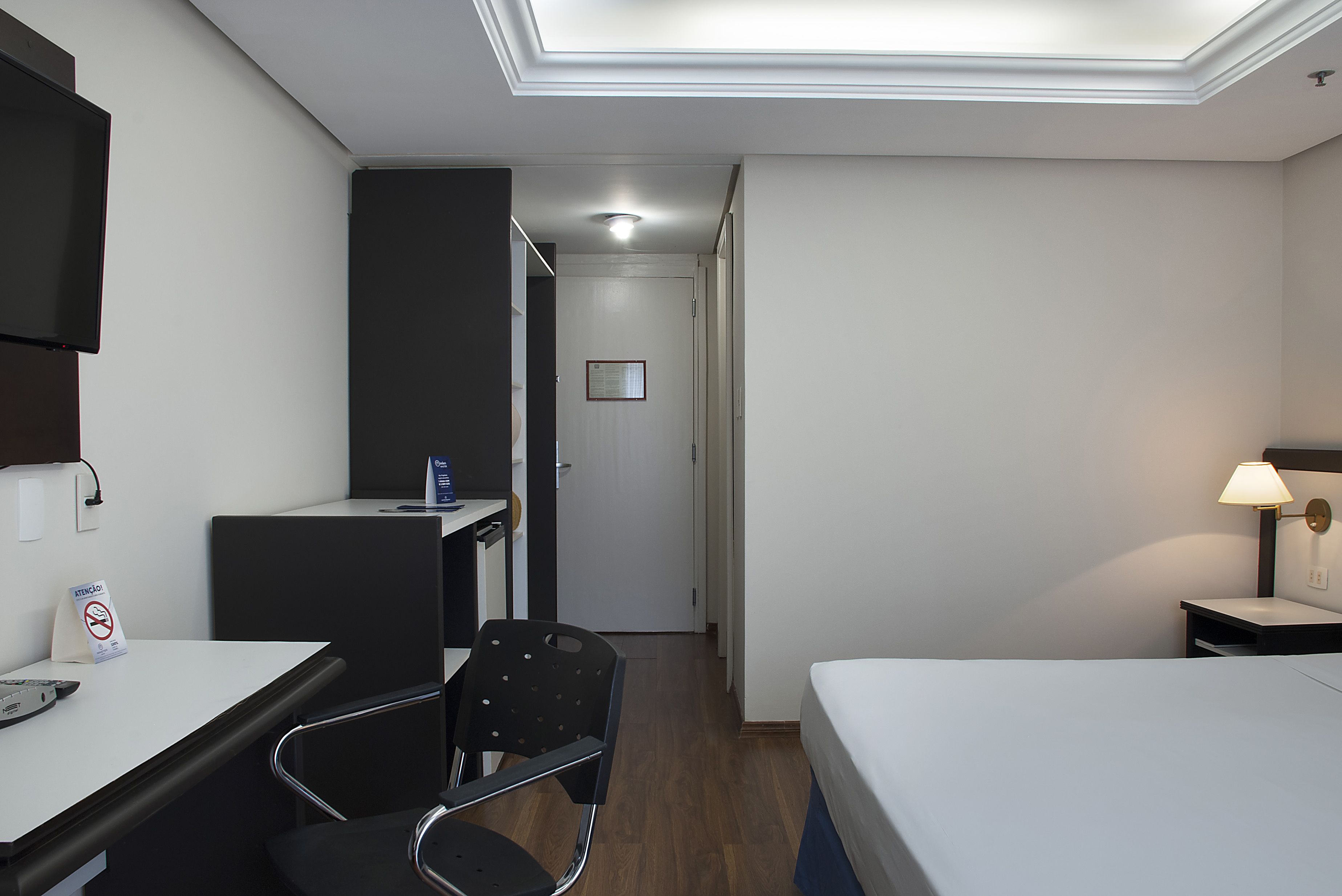 A hotel room with a bed, desk, chair, and a small table with a lamp.