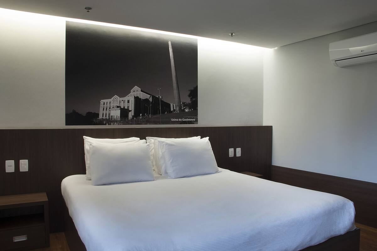 A hotel room with a large bed, a nightstand, a wall-mounted air conditioner, and a poster of a building.