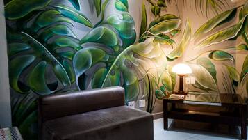 Habitación con un gran mural de hojas tropicales, un sofá marrón y una pequeña mesa con lámpara.