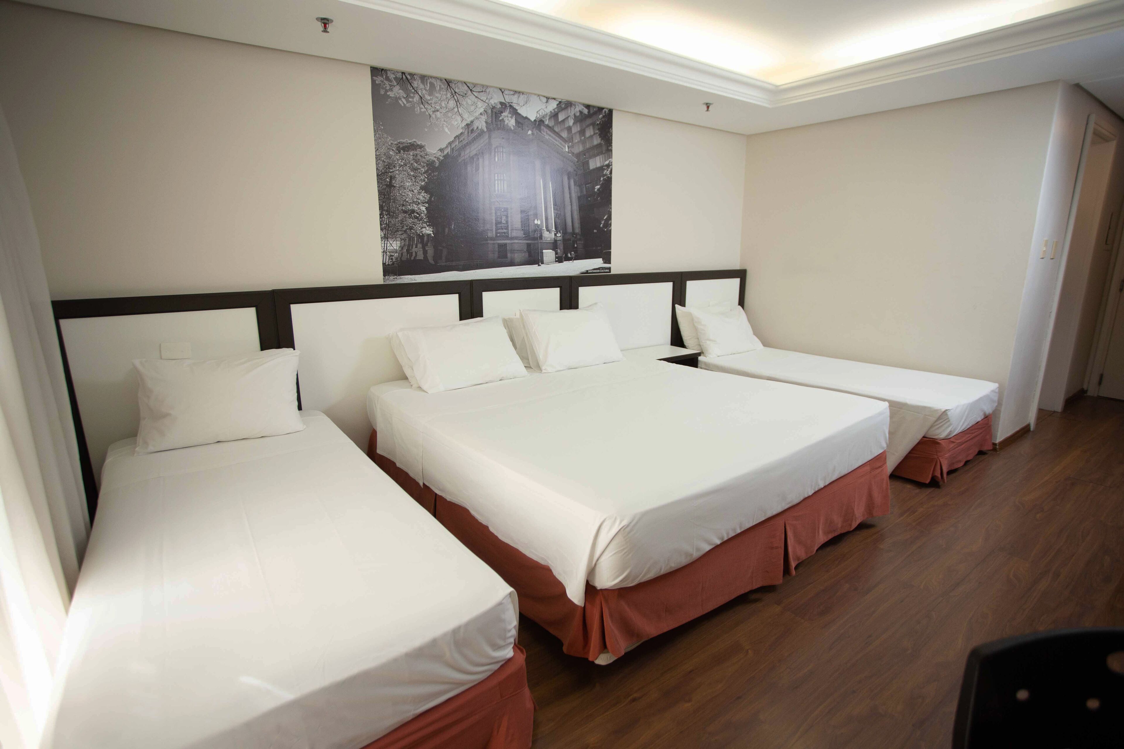 Una habitación de hotel con tres camas dispuestas en línea, un cuadro grande enmarcado en la pared y suelo de madera.
