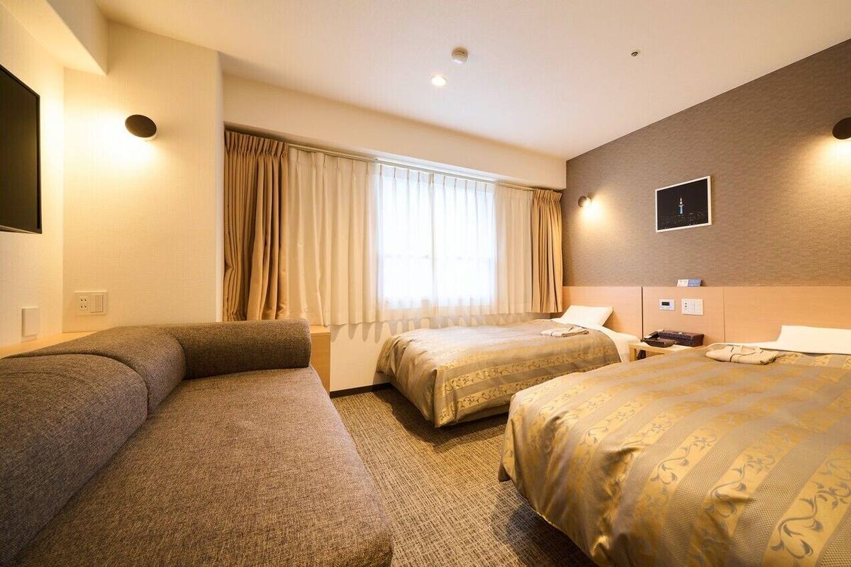Ein Hotelzimmer mit zwei Betten, einem großen Fenster mit Vorhängen, einem Flachbildfernseher und einer wandmontierten Bedieneinheit.