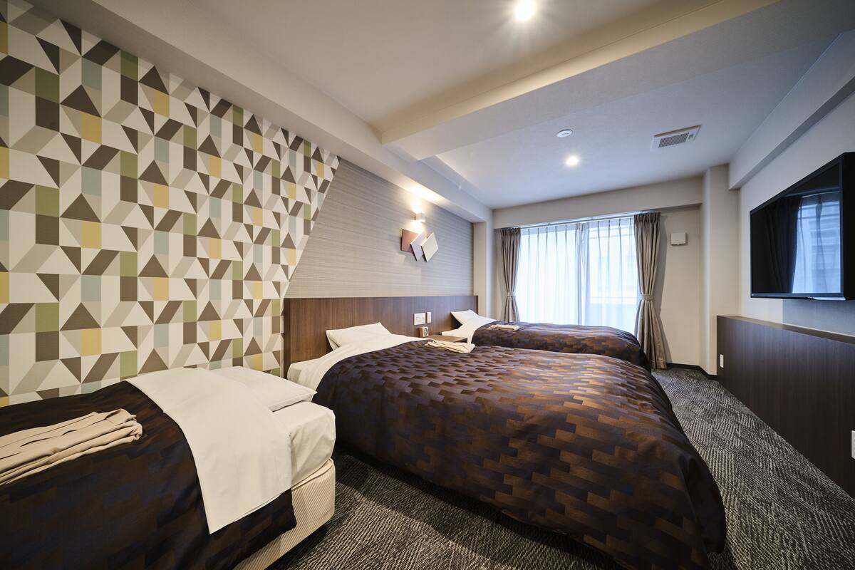 Ein Hotelzimmer mit zwei Betten, einer Wand mit Muster, einem Flachbildfernseher und einem Fenster mit Vorhängen.