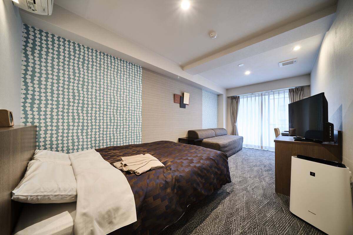 Ein Hotelzimmer mit einem großen Bett, einem Schreibtisch mit Fernseher, einem Sessel und einem kleinen Kühlschrank.