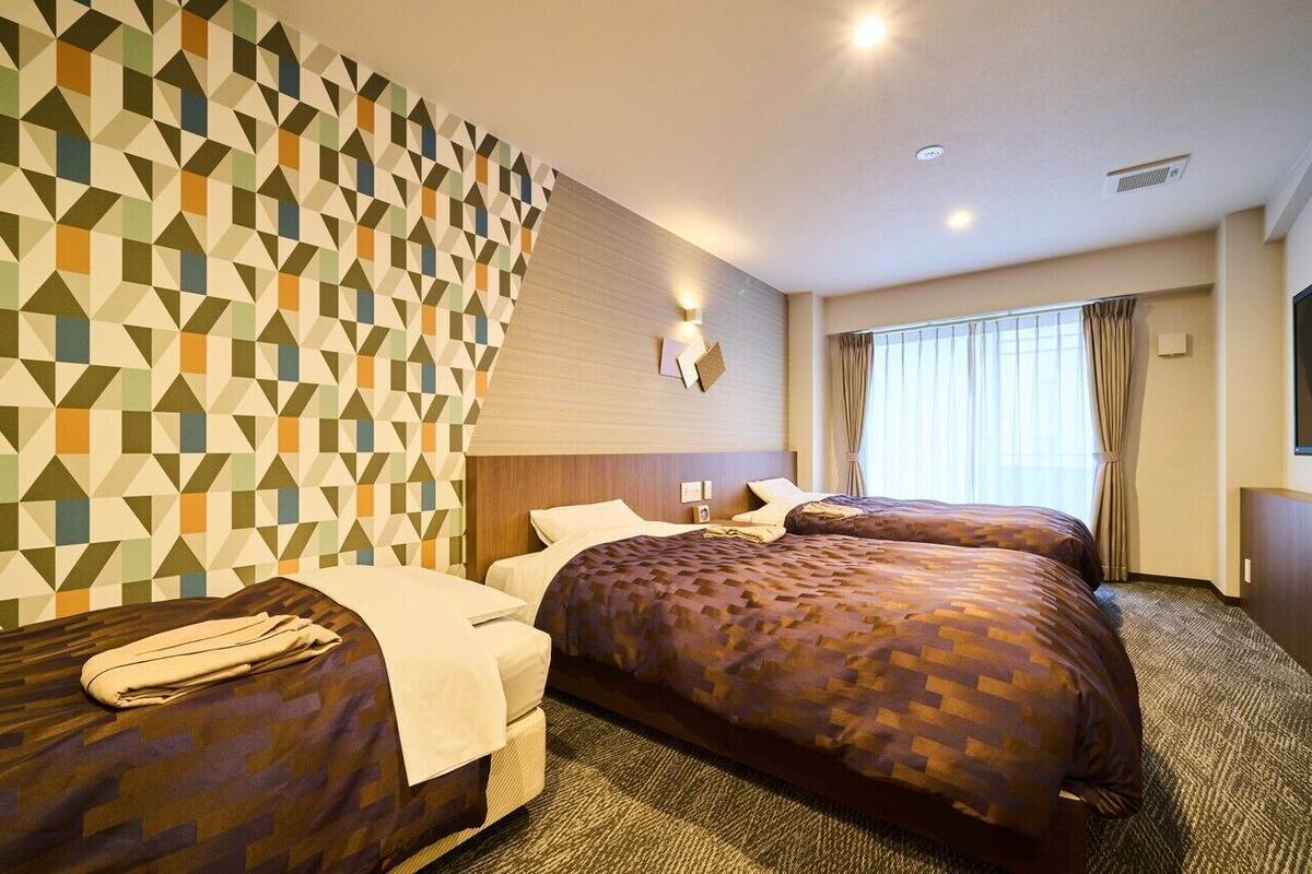 Ein Hotelzimmer mit zwei Betten, einer Wand mit geometrischem Muster, einem Flachbildfernseher und einem Fenster mit Vorhängen.