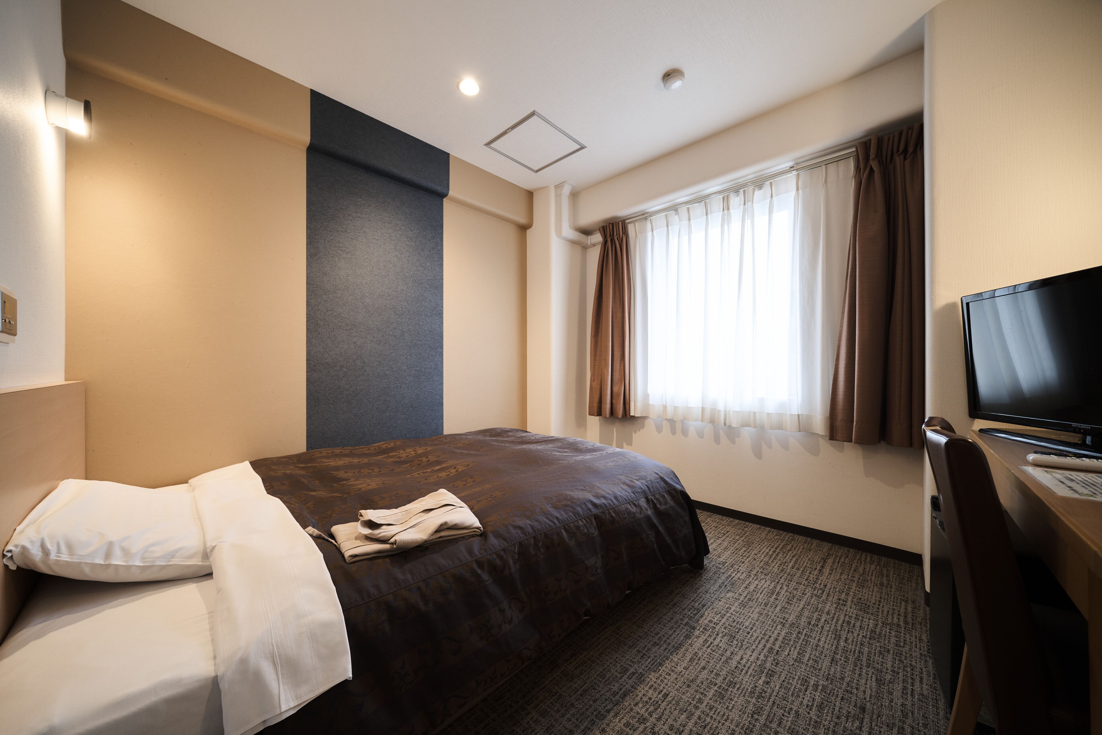 Ein Hotelzimmer mit einem Bett, einem Schreibtisch mit Computer, einem Fernseher und einem Fenster mit Vorhängen.