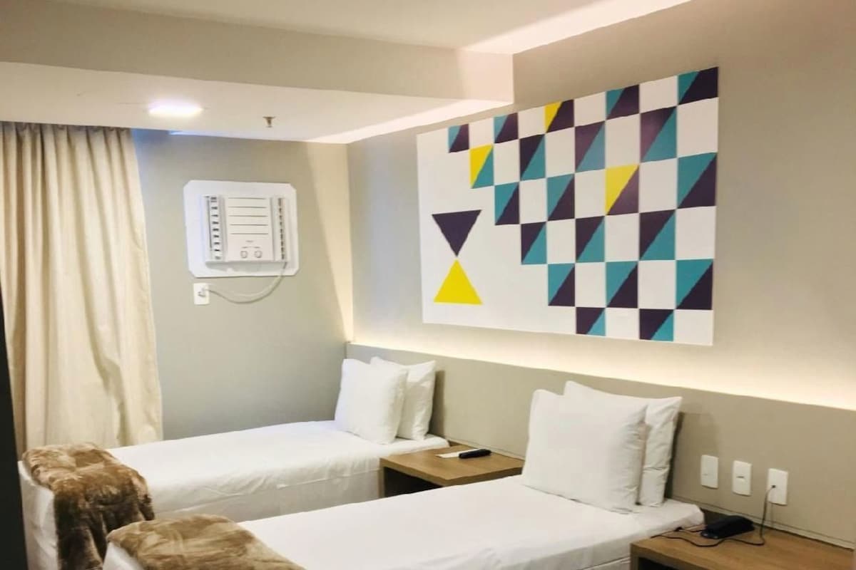 A hotel room with two beds, a large abstract painting on the wall, a bedside table with a lamp, and a power outlet.