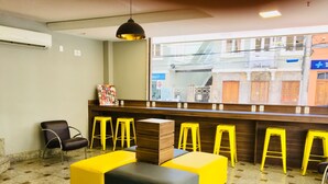 Un hall d’entrée avec un comptoir de réception, des tabourets jaunes, un espace salon aux couleurs noir et jaune, et une grande fenêtre donnant sur des immeubles.
