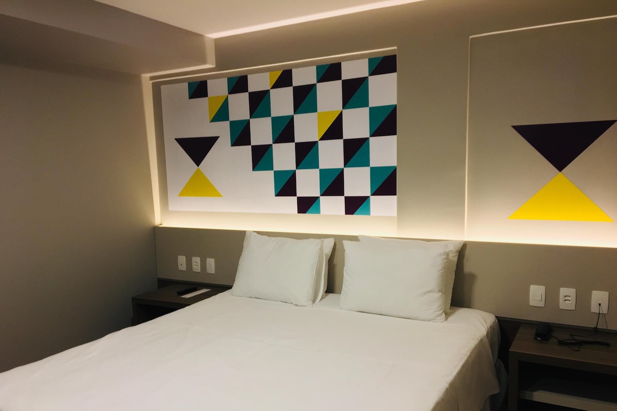 A hotel room with a bed, two bedside tables, and a wall with geometric artwork.
