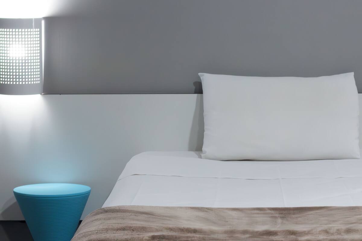 A neatly made bed with a white mattress and a brown blanket, a white pillow, and a blue nightstand with a lamp.