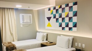 Une chambre d’hôtel avec deux lits, un climatiseur fixé au mur, une table de chevet avec une télécommande et une œuvre d’art moderne aux motifs géométriques accrochée au mur.