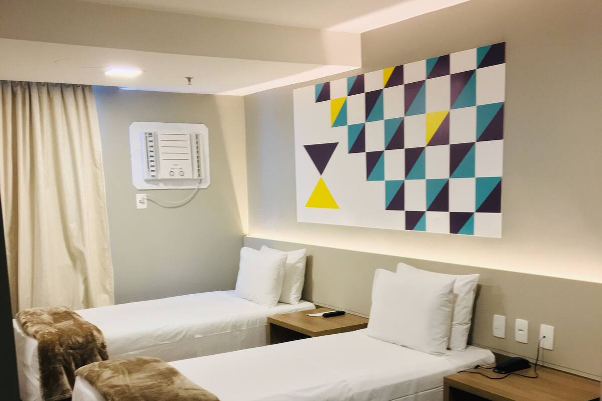 A hotel room with two beds, a wall-mounted air conditioning unit, a bedside table with a remote control, and a modern geometric wall art piece.