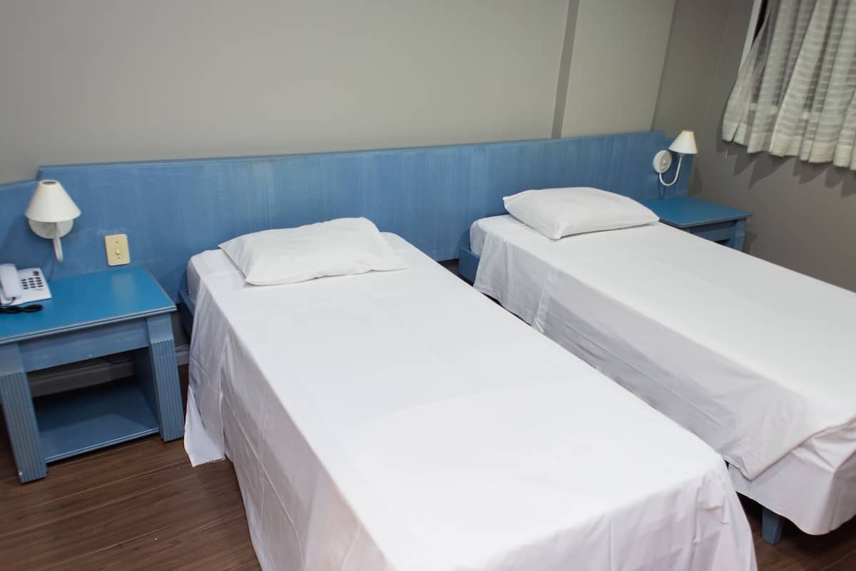 A hotel room with two single beds, a blue nightstand with a lamp, and a phone.
