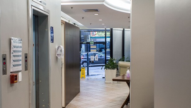 A hotel lobby with a tiled floor, an elevator, a trash bin, a reception desk, and a seating area with a plant. - Master Express Alberto Bins (Porto Alegre)