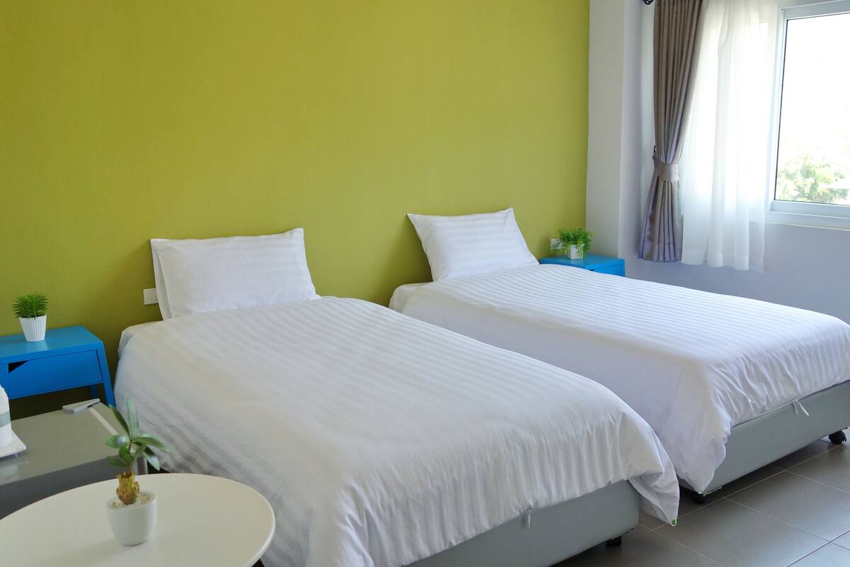 A hotel room with two beds, a small table with a plant, a blue nightstand, and a window with curtains.