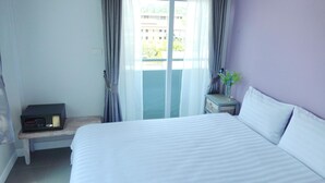 Camera d'albergo con un letto, un tavolino con una radio, un comodino con un vaso di fiori e vista sugli edifici attraverso la finestra.