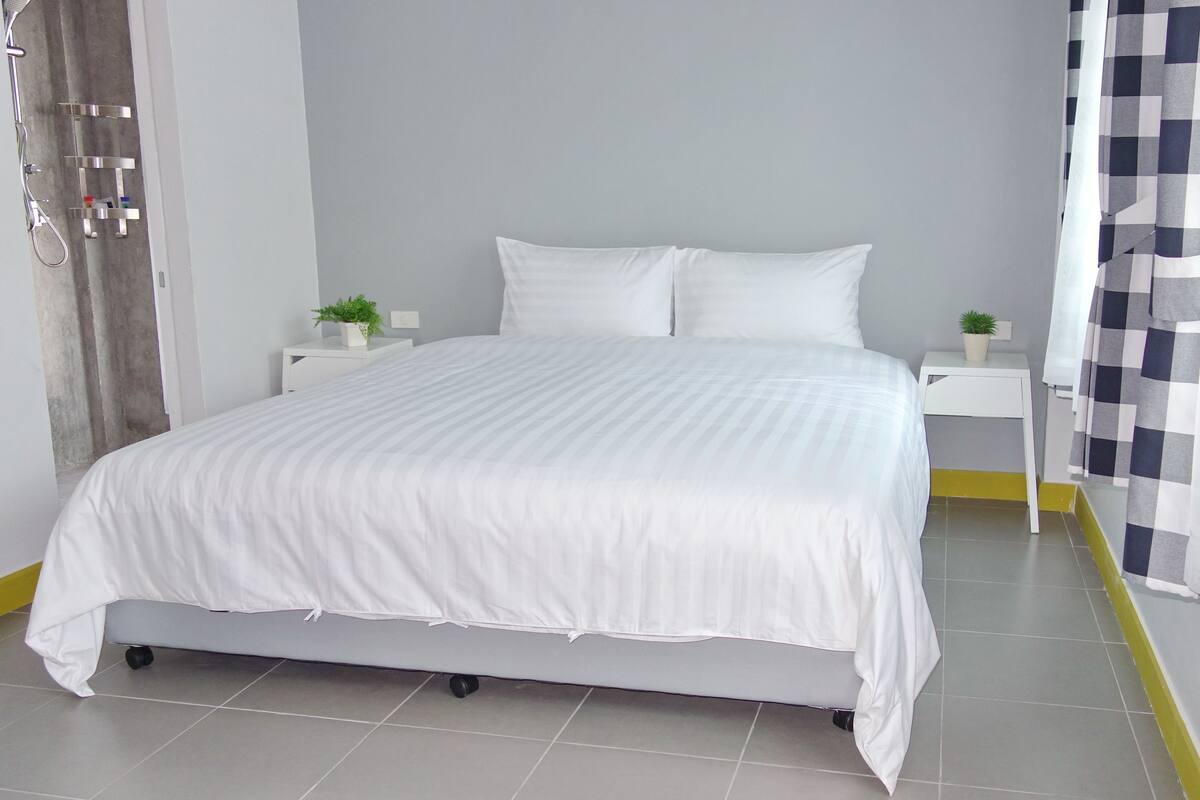 A neatly made bed with white bedding, a small bedside table with a plant, and a checkered curtain.