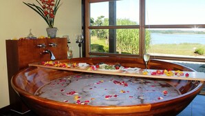 A large wooden bathtub filled with water and rose petals, a wooden vanity with a sink and a potted plant, and a view of a lake through a large window. - Balmer See - Hotel · Golf · Spa (Benz)