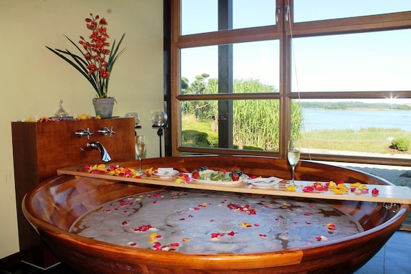 Eine große Holzwanne, gefüllt mit Wasser und Rosenblüten, ein Holzbadezimmermöbel mit Waschbecken und einer Topfpflanze, und Blick auf einen See durch ein großes Fenster.