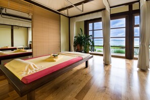 A room with wooden floors, a massage table, a window with a view of the sea, and a potted plant. - Balmer See - Hotel · Golf · Spa (Benz)