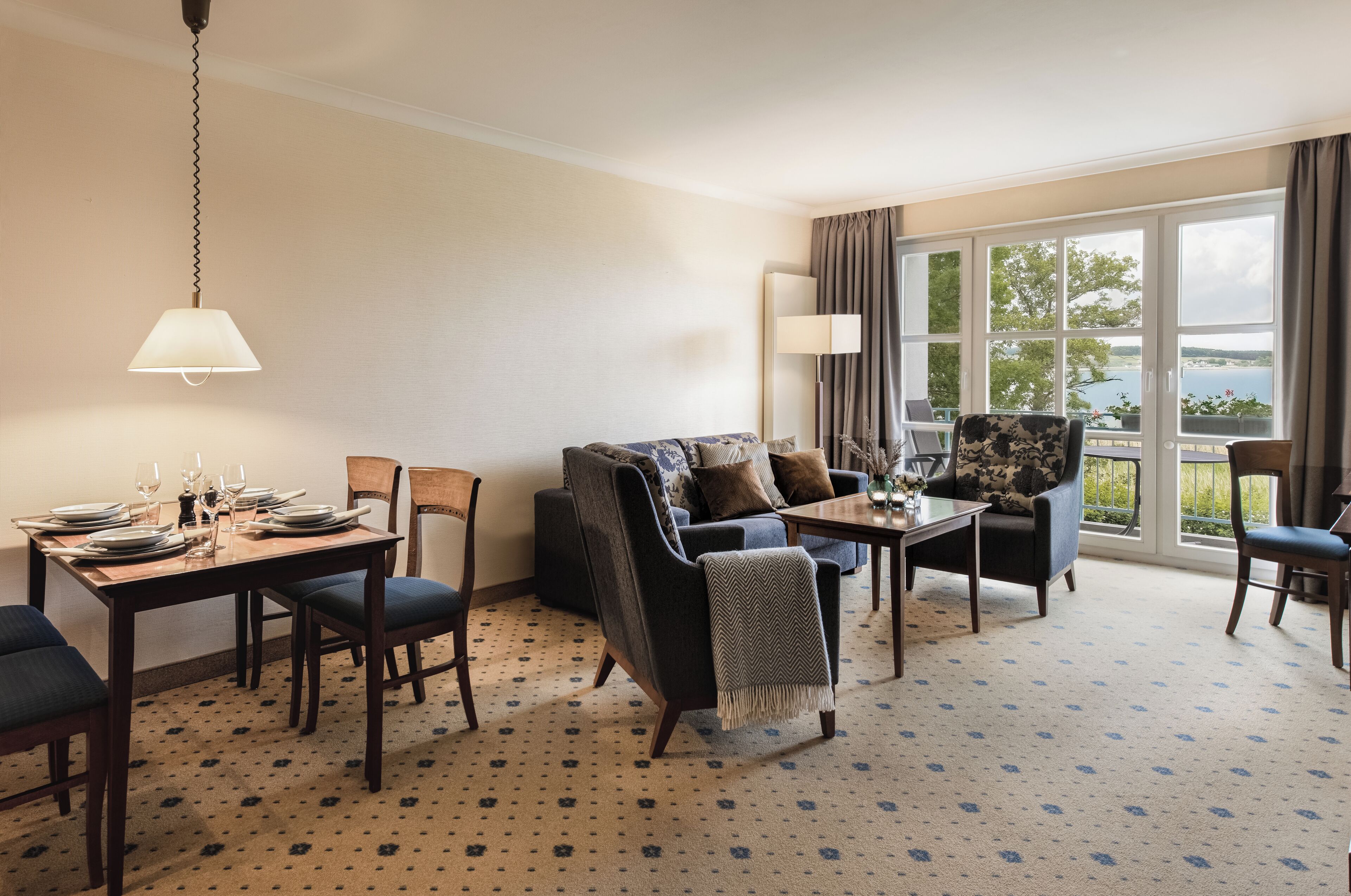 A well-lit room with a dining table set for four, a sofa, and a chair. A large window provides a view of the outdoors.