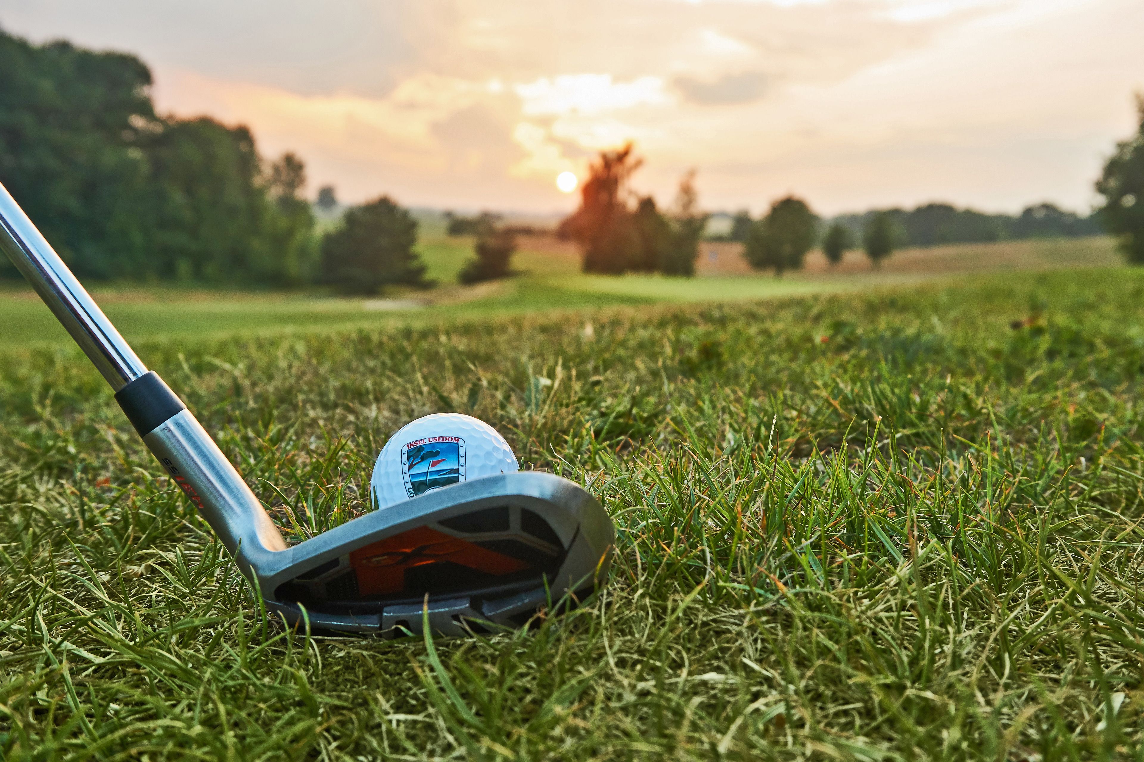 Un palo de golf sobre un campo cubierto de césped, una pelota de golf y una puesta de sol al fondo.