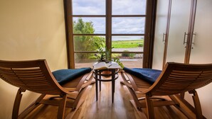 A room with a wooden table and two chairs with blue cushions, a window with a view of greenery and a body of water, and a wooden cabinet to the right. - Balmer See - Hotel · Golf · Spa (Benz)