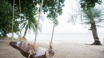 A wooden swing suspended by ropes, overlooking a sandy beach with palm trees and a view of the sea.