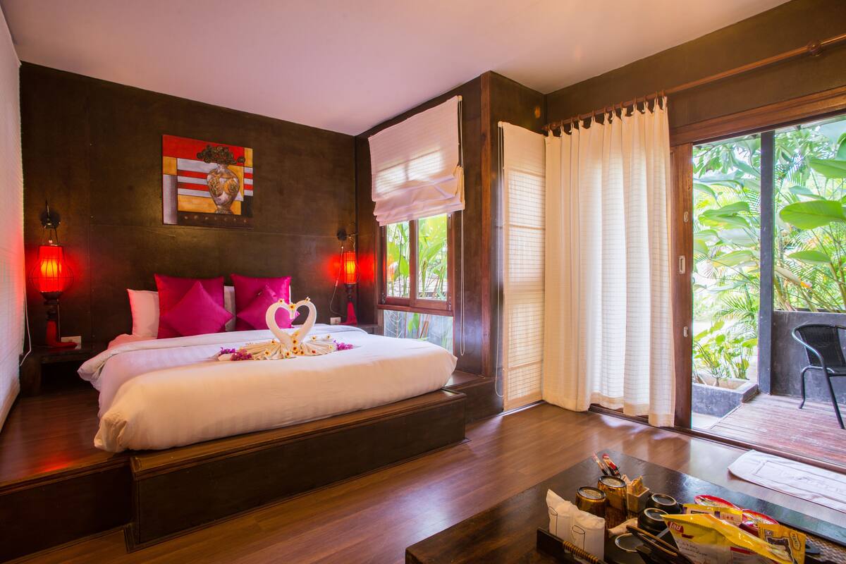 A bedroom with a bed, pillows, a nightstand, a window with curtains, and a view of greenery outside.